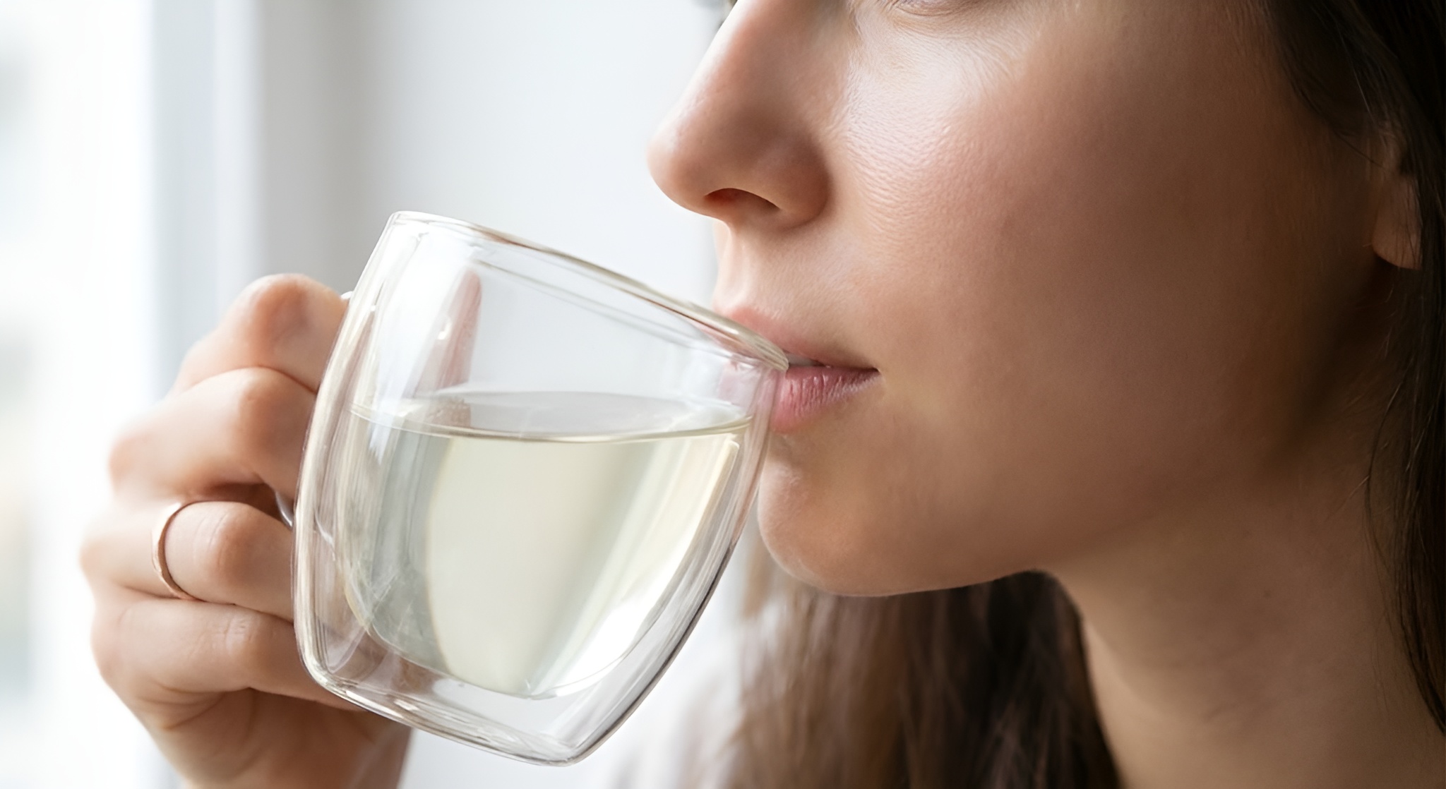 A close up of a womans flawless skin as she sips a white tea in a clear glass mug.